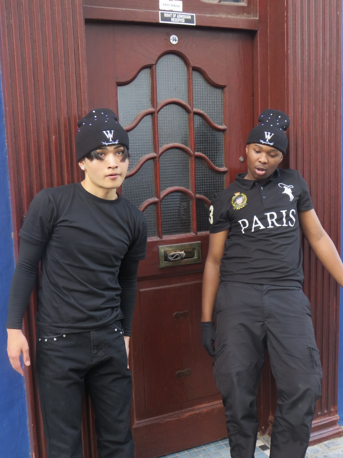 Two people wearing black WANOFWAN beanies in front of a red doorway